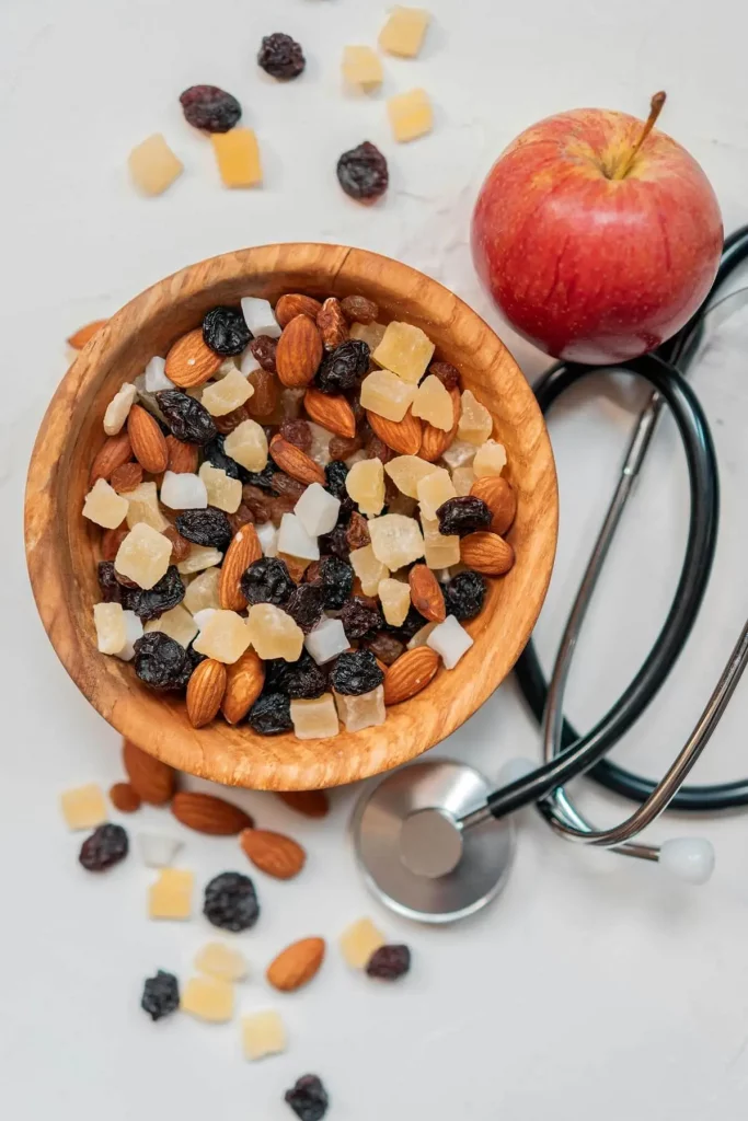 Un bol en bois rempli de noix, de fruits secs et de noix de coco, à côté d'une pomme rouge et d'un stéthoscope sur une surface blanche.